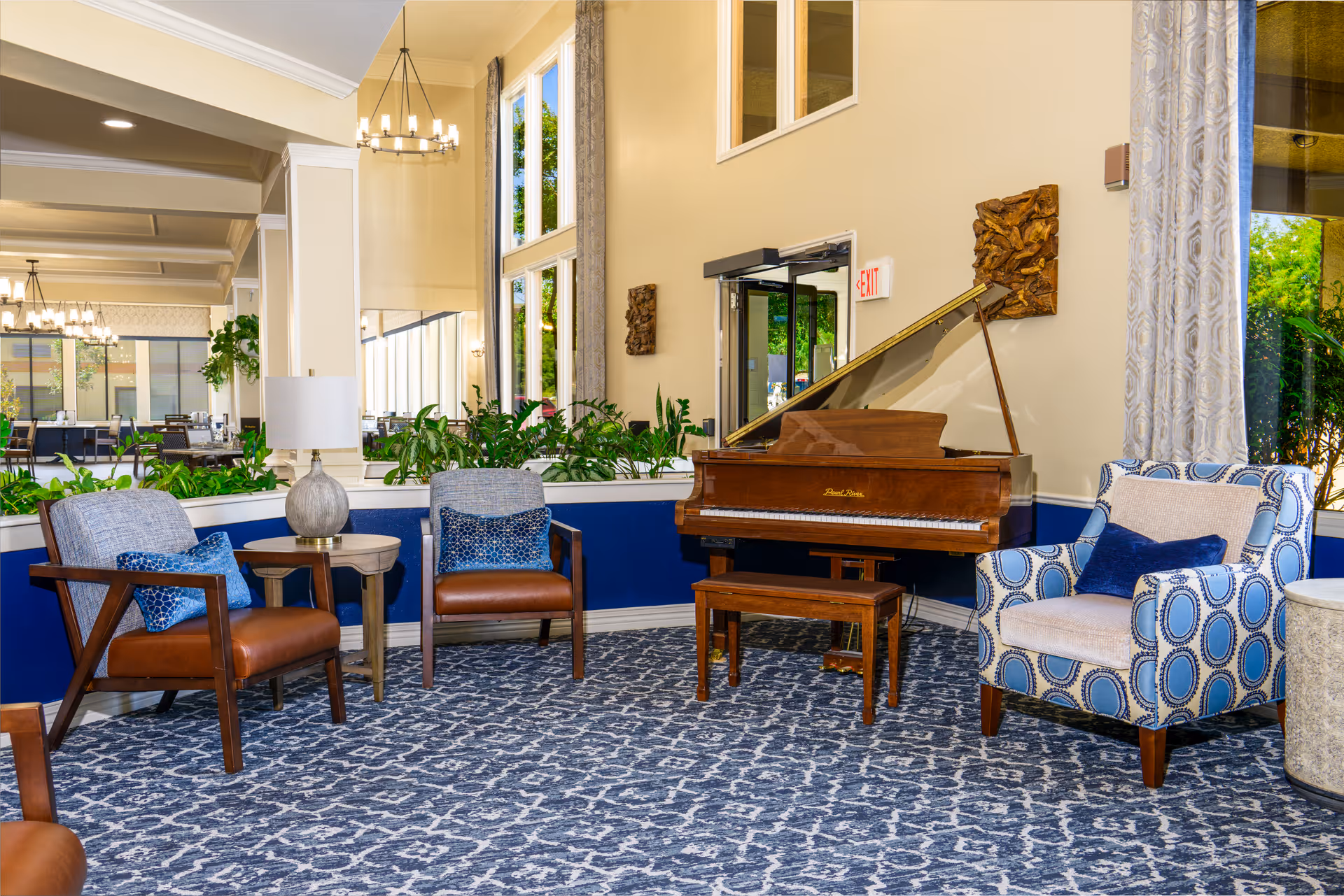A cozy seating area in a senior living facility with a grand piano, two brown leather chairs with blue patterned pillows, a blue and white patterned armchair with a blue pillow, a small round wooden table with a lamp, and large windows letting in natural light. The room has a blue and white patterned carpet and beige walls with decorative wall art.