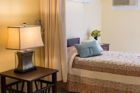 A cozy bedroom with a bed covered in a floral quilt and two pillows, one blue and one white. Next to the bed is a dark wooden nightstand with a decorative table lamp. A beige curtain hangs beside the bed, and a small window air conditioning unit is visible on the wall.