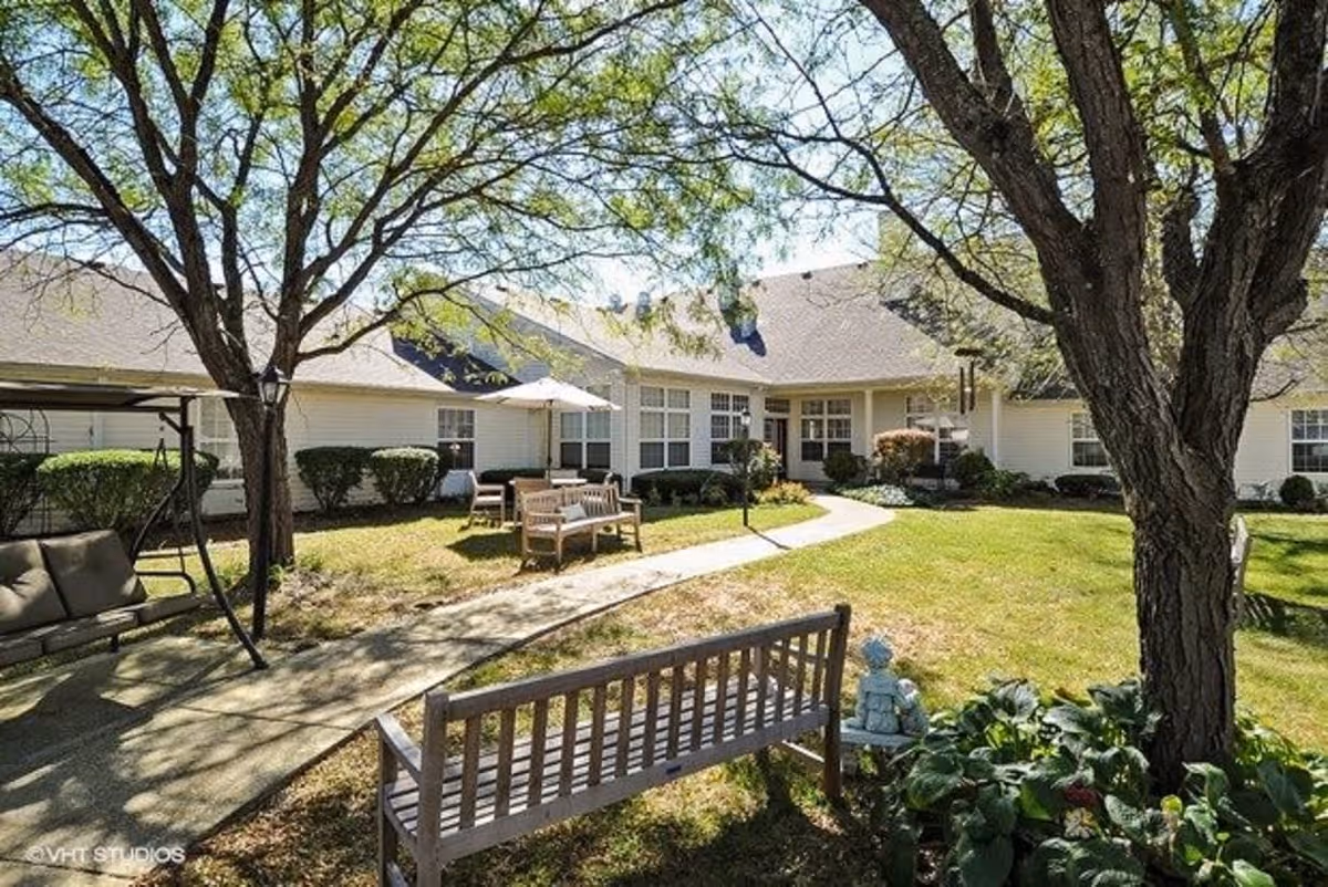 Outdoor courtyard area at Keepsake Village of Columbus Memory Care featuring a paved walkway, wooden benches, a swing seat, trees, and a building with multiple windows in the background under a clear sky.