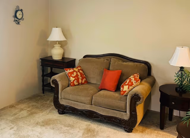 A cozy living room corner featuring a brown upholstered loveseat with carved wooden trim, adorned with three decorative pillows in red and patterned designs. On either side of the loveseat are dark wooden side tables, each with a white table lamp. A decorative wall clock is mounted on the beige wall above the left side table, and a green plant is partially visible on the right side near the other table.