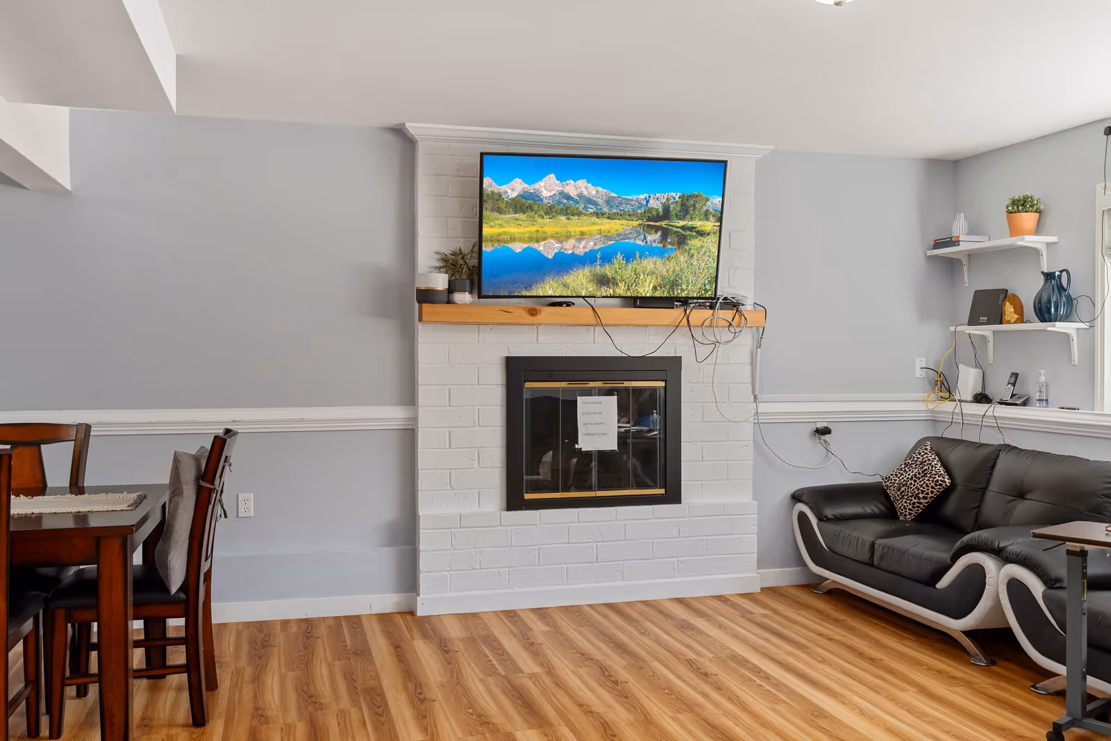 A cozy living room with light gray walls and wood flooring. There is a white brick fireplace with a wooden mantel, above which a flat-screen TV displays a scenic mountain and lake view. To the right of the fireplace is a black and white modern sofa with a leopard print pillow. On the left side, there is a wooden dining table with chairs. Shelves with decorative items and plants are mounted on the wall near the sofa.