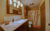 A bathroom with a granite countertop vanity featuring a sink, soap dispenser, and decorative items. Above the vanity is a large mirror with four hanging light fixtures. The room has a toilet next to a bathtub with a patterned shower curtain. The floor is wooden, and there is a red bath mat in front of the tub.