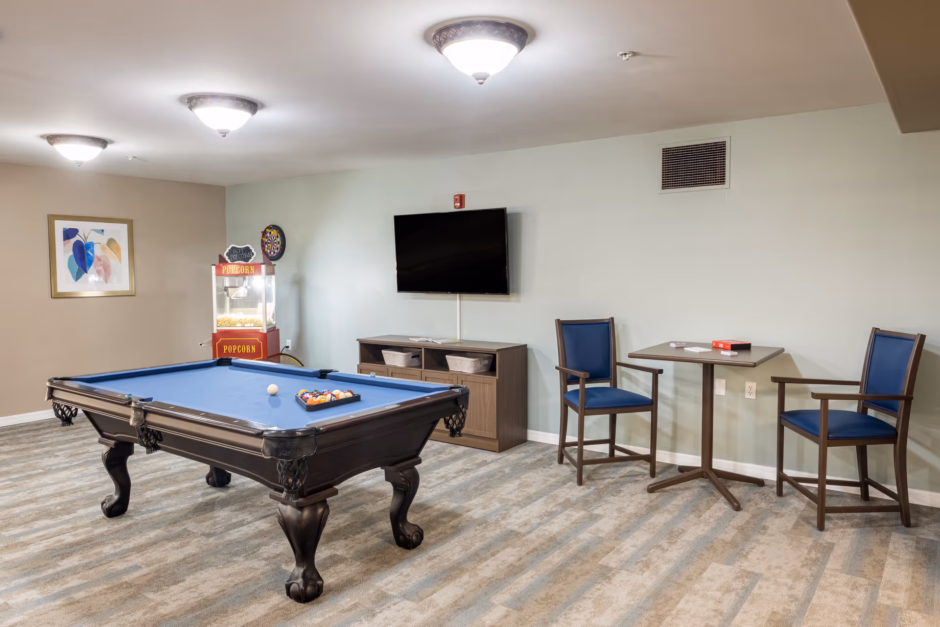 A recreational room with a blue pool table in the center, a popcorn machine in the corner, a wall-mounted TV above a wooden cabinet, and a small table with two blue cushioned chairs. The room has light-colored walls and carpeted flooring.
