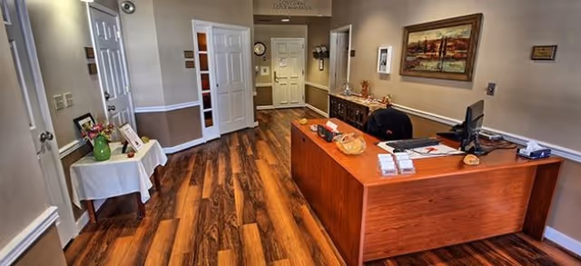 Reception area in a senior living facility with a wooden desk, computer, and office chair. The room has wood flooring, beige walls with white trim, a small table with flowers and framed photos, and a painting on the wall. Several doors lead to other rooms.