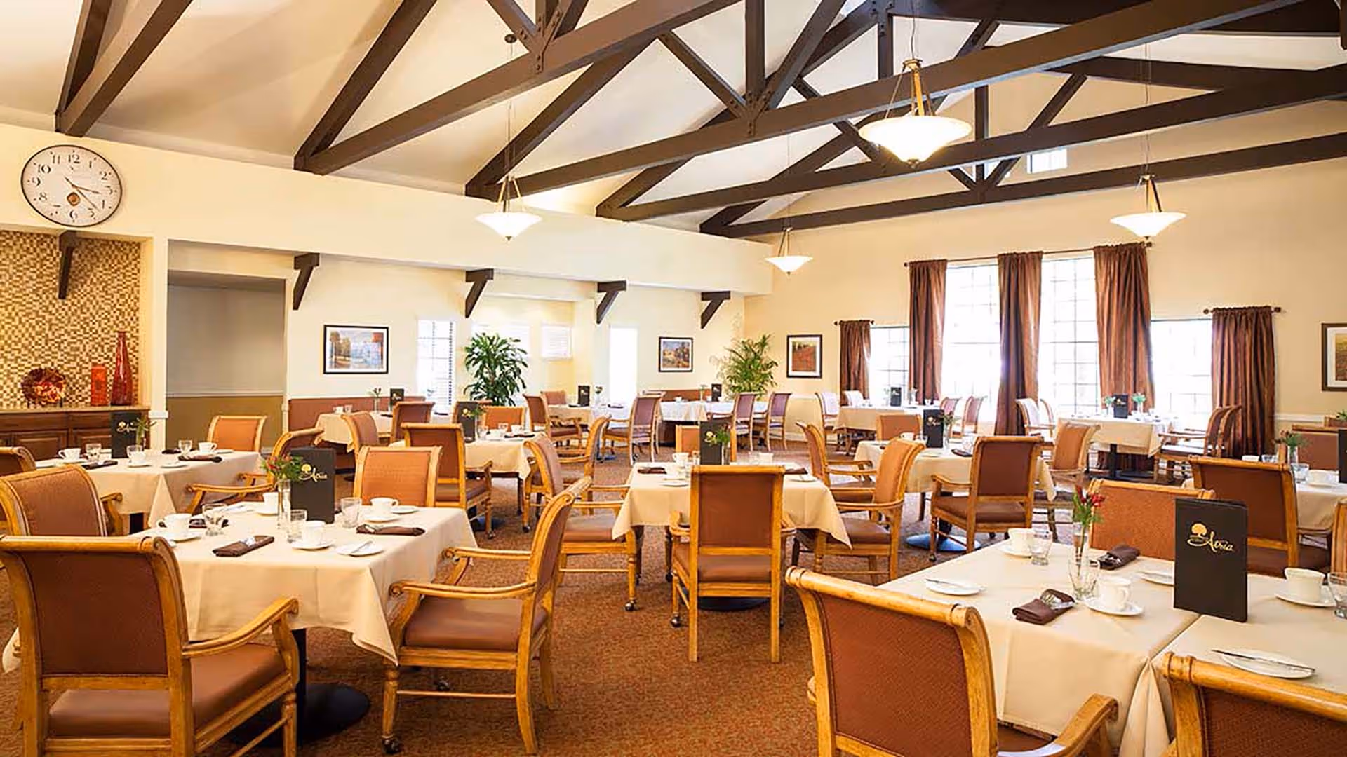 A spacious dining room with multiple tables covered in white tablecloths, each set with cups, plates, and menus. The room features high ceilings with exposed wooden beams, large windows with brown curtains, and several potted plants. The chairs are wooden with cushioned seats and backs.