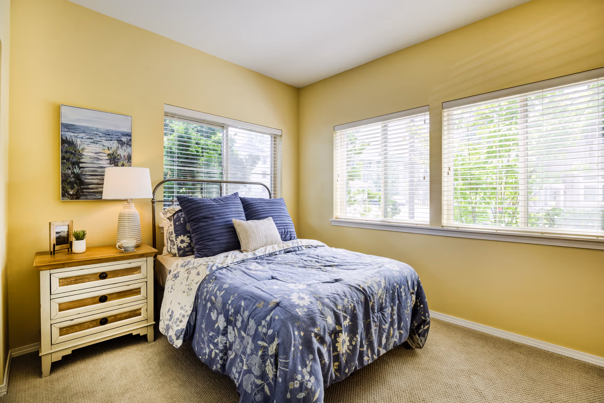 Sunlit bedroom with a bed dressed in blue floral bedding, a nightstand with a lamp and artwork, and three large windows with blinds.