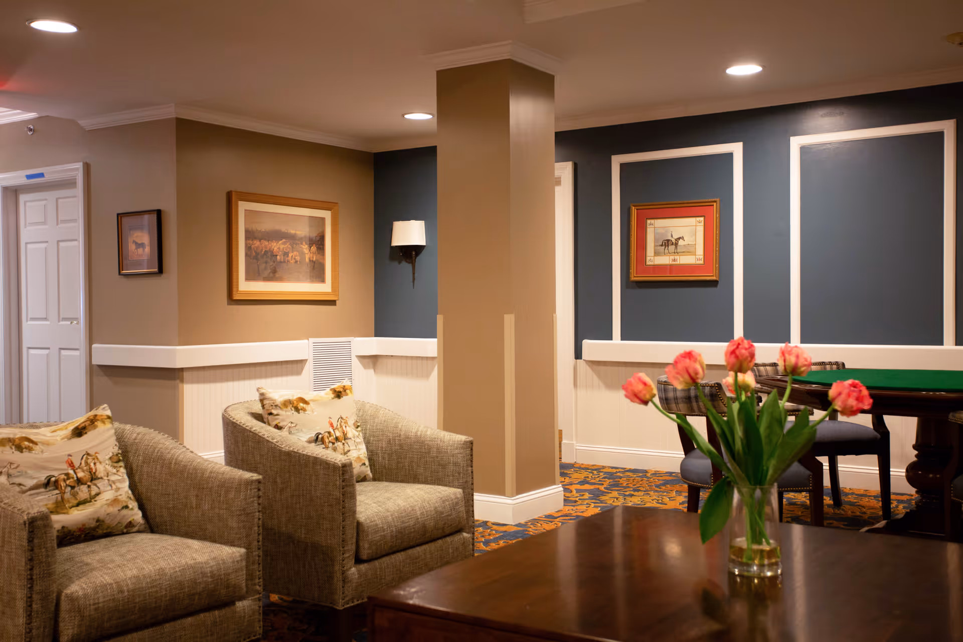 A cozy interior lounge area with two beige armchairs featuring horse-themed cushions, a wooden table with a vase of pink tulips, framed horse artwork on the walls, and a green felt card table with chairs in the background. The walls are painted in beige and dark blue with white trim, and the carpet has a decorative pattern.