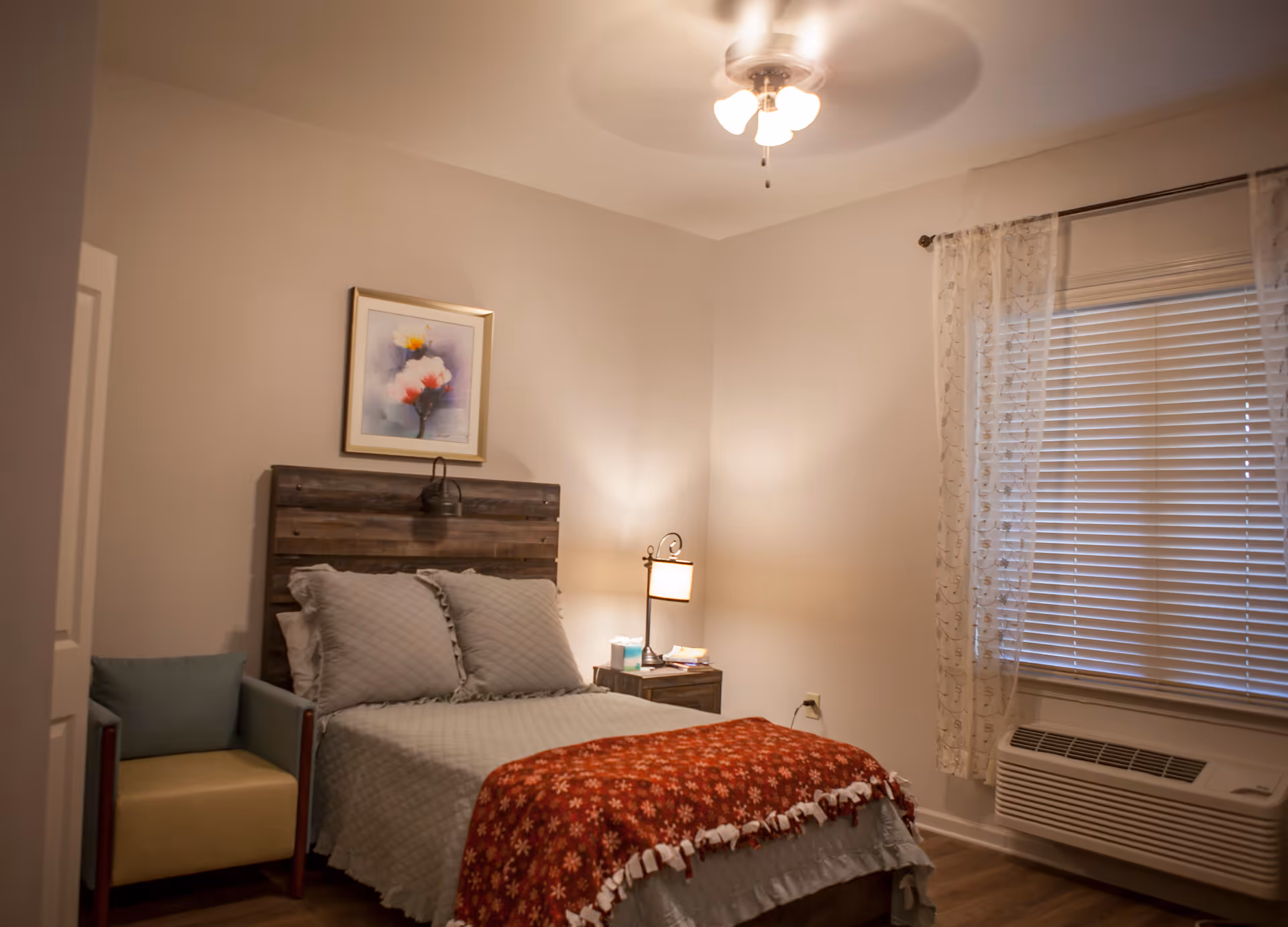 Bedroom with a made bed featuring a wooden headboard, bedside lamp, armchair, ceiling fan, window blinds and an air conditioner.