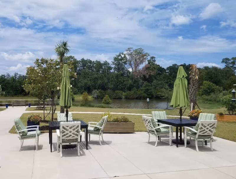 Outdoor patio area with two black tables, each surrounded by four white chairs with green and white striped cushions. Each table has a closed green umbrella. The patio overlooks a grassy area with trees and a body of water under a partly cloudy sky.