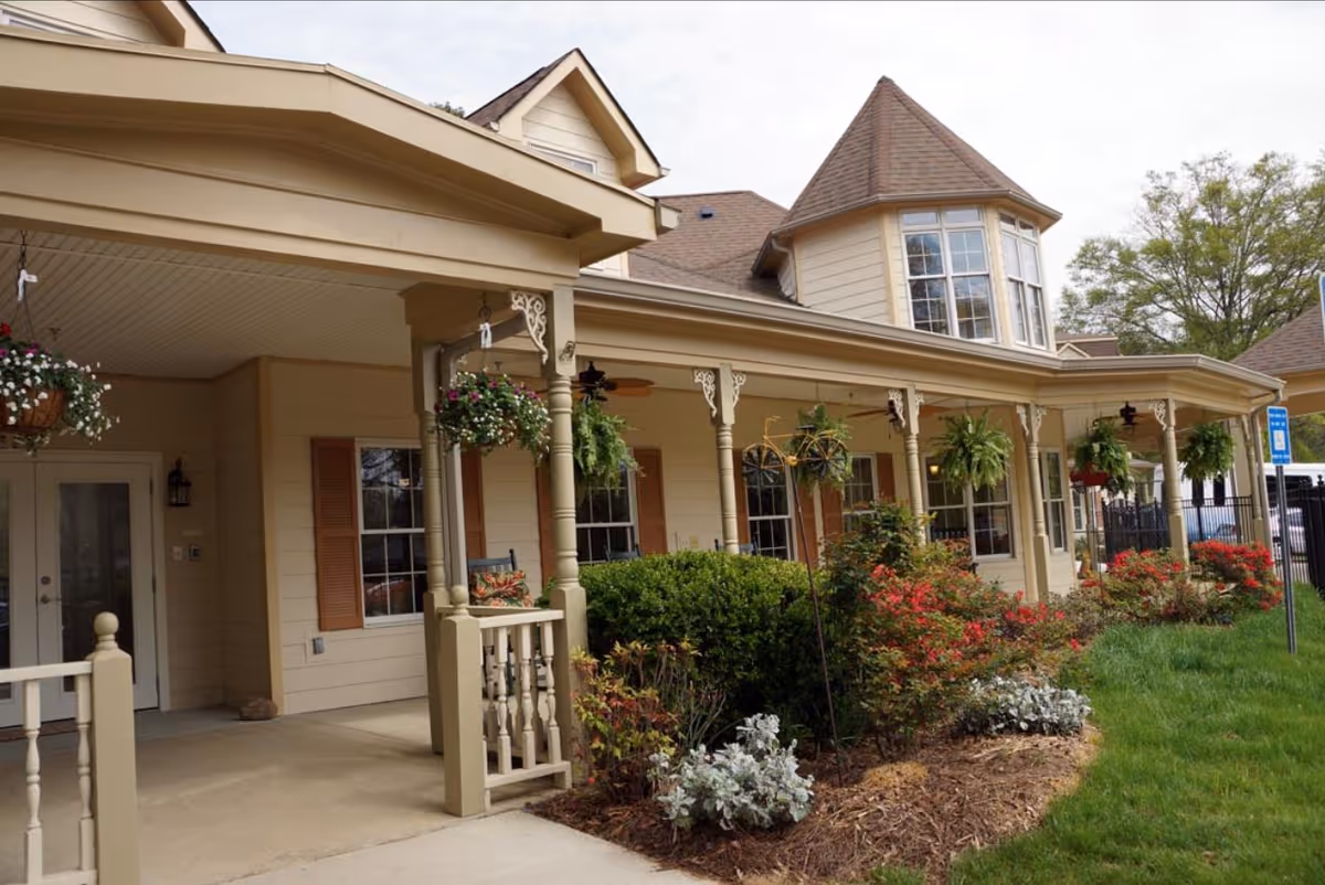 Exterior view of a senior living facility with a covered porch featuring hanging flower baskets and decorative columns. The building has beige siding, multiple windows with brown shutters, and a turret-like architectural feature. There are well-maintained bushes and flowering plants along the front, with a grassy area and a parking sign visible.