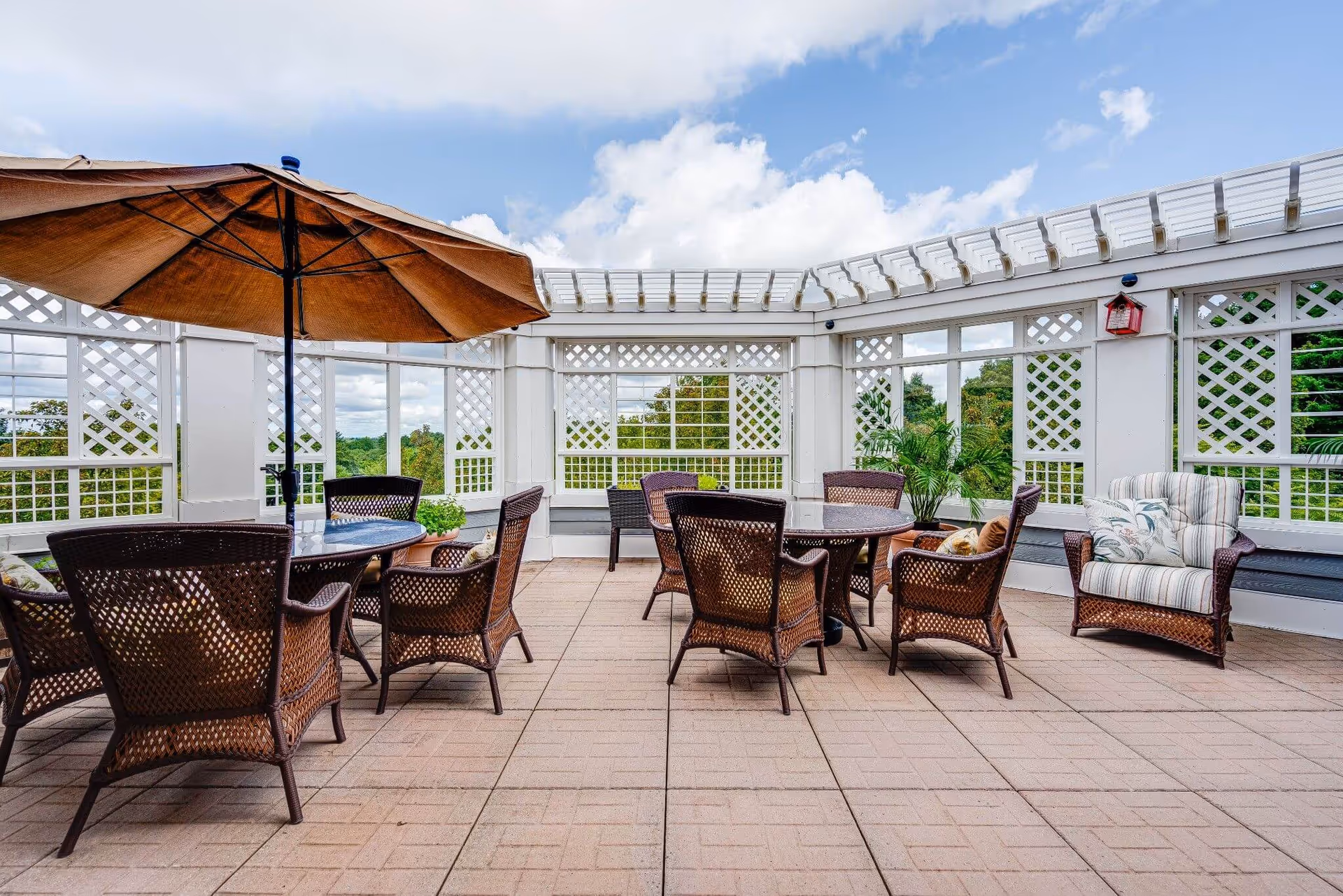 Open-air rooftop patio with wicker tables and chairs, a large umbrella, lattice pergola, and potted plants overlooking greenery.