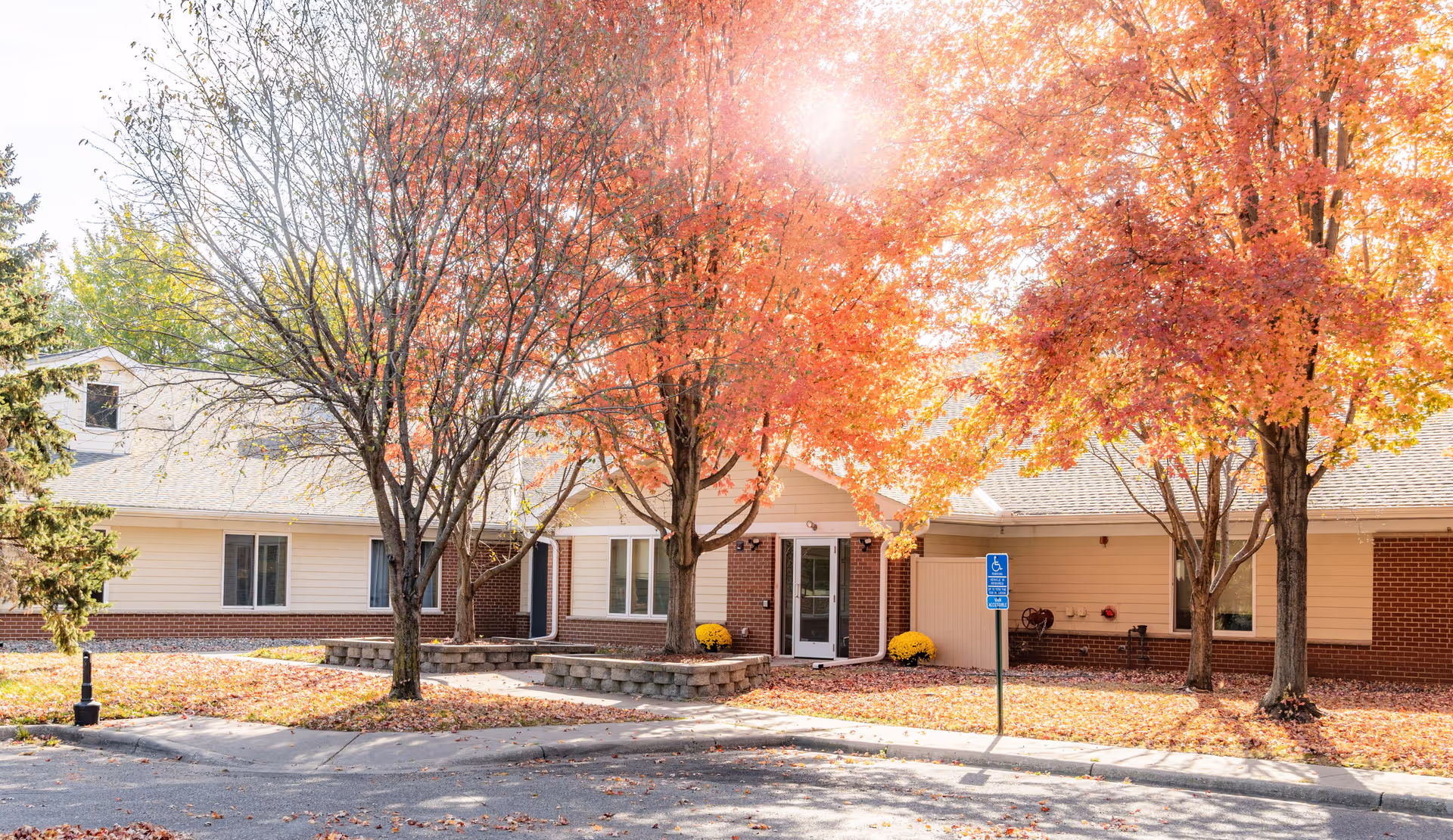 Exterior view of Southern Oaks Place facility during autumn with trees displaying vibrant orange and red leaves, a brick and beige siding building, a sidewalk, and a handicapped parking sign in front.