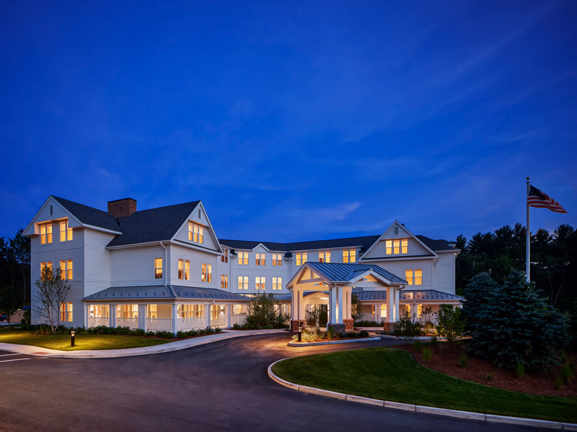 Exterior view of Stone Hill at Andover senior living facility at dusk, showing a large white multi-story building with many lit windows, a covered entrance, landscaped greenery, and an American flag on a flagpole.