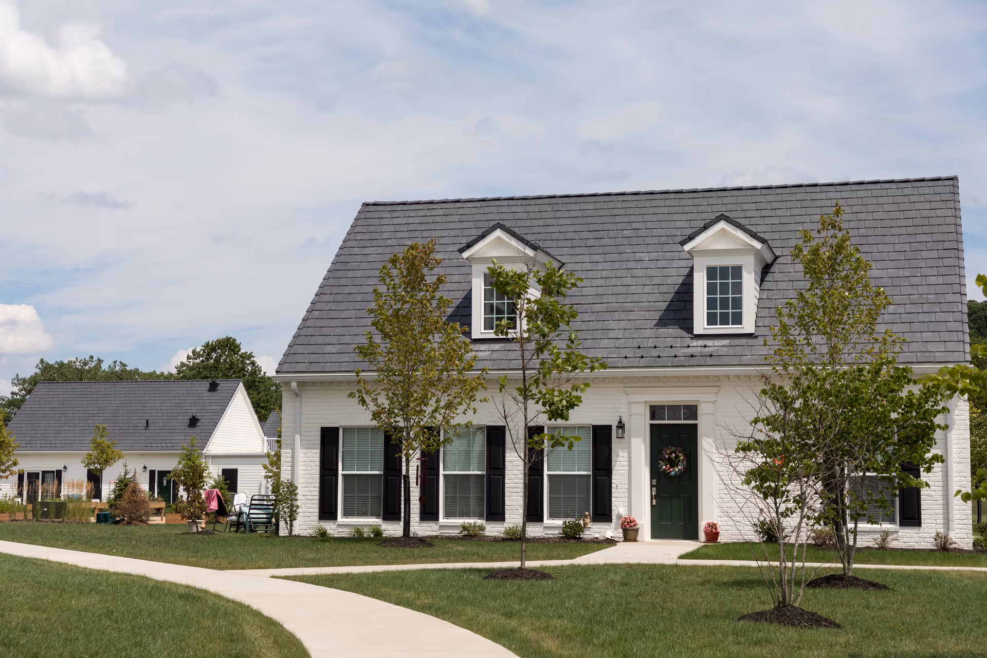A white brick residential building with a gray shingled roof and two dormer windows. The front door is green with a decorative wreath, flanked by black shutters on the windows. There are young trees and neatly maintained grass in the front yard, with a curved concrete walkway leading to the entrance. Another similar building is visible in the background under a partly cloudy sky.