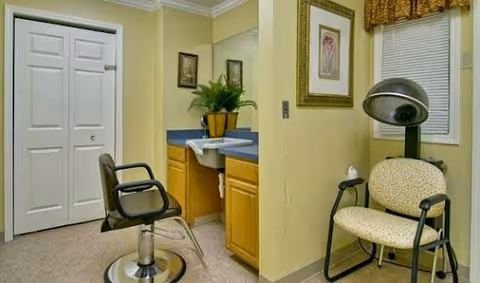 Interior view of a senior living facility room with a salon chair in front of a sink and counter, a drying hood chair, a closet with double doors, and framed artwork on the walls.