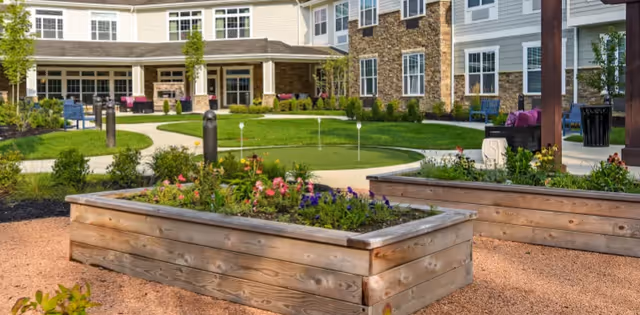 Outdoor garden area with raised wooden flower beds filled with colorful flowers, a putting green with golf holes, and a building with large windows and a covered patio in the background.