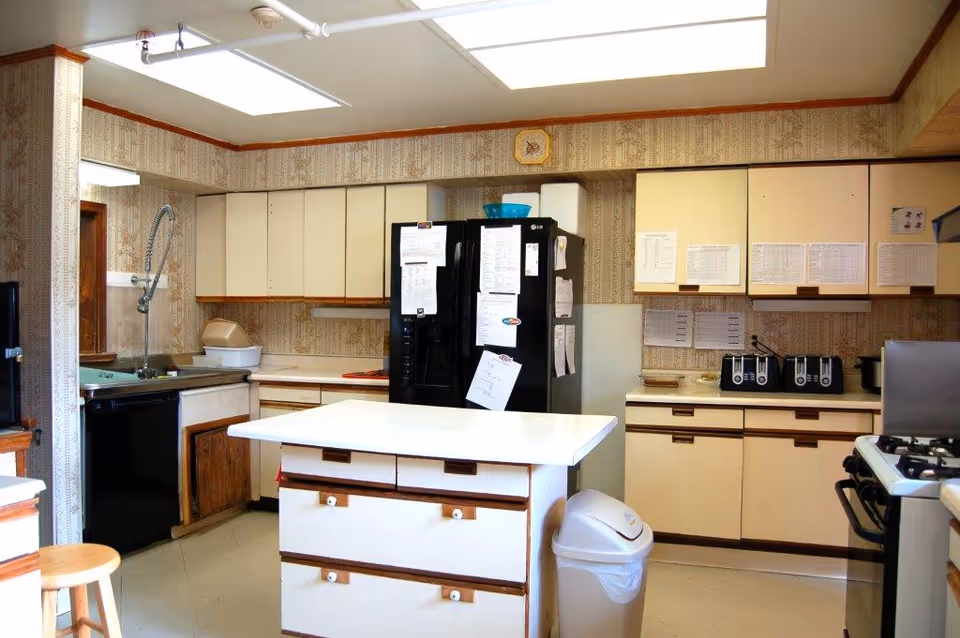 Well-lit institutional kitchen with a central island, refrigerator, stove, sink, and cabinets.