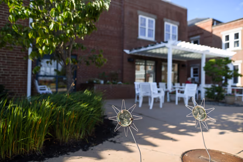 Outdoor patio area at a senior living facility with white chairs and tables under a white pergola, surrounded by greenery and brick building walls. Decorative sun-shaped garden stakes are visible in the foreground.