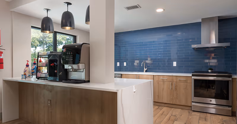 Modern communal kitchen with a white island, coffee machines, blue tiled backsplash, stainless steel stove, and wooden cabinets.