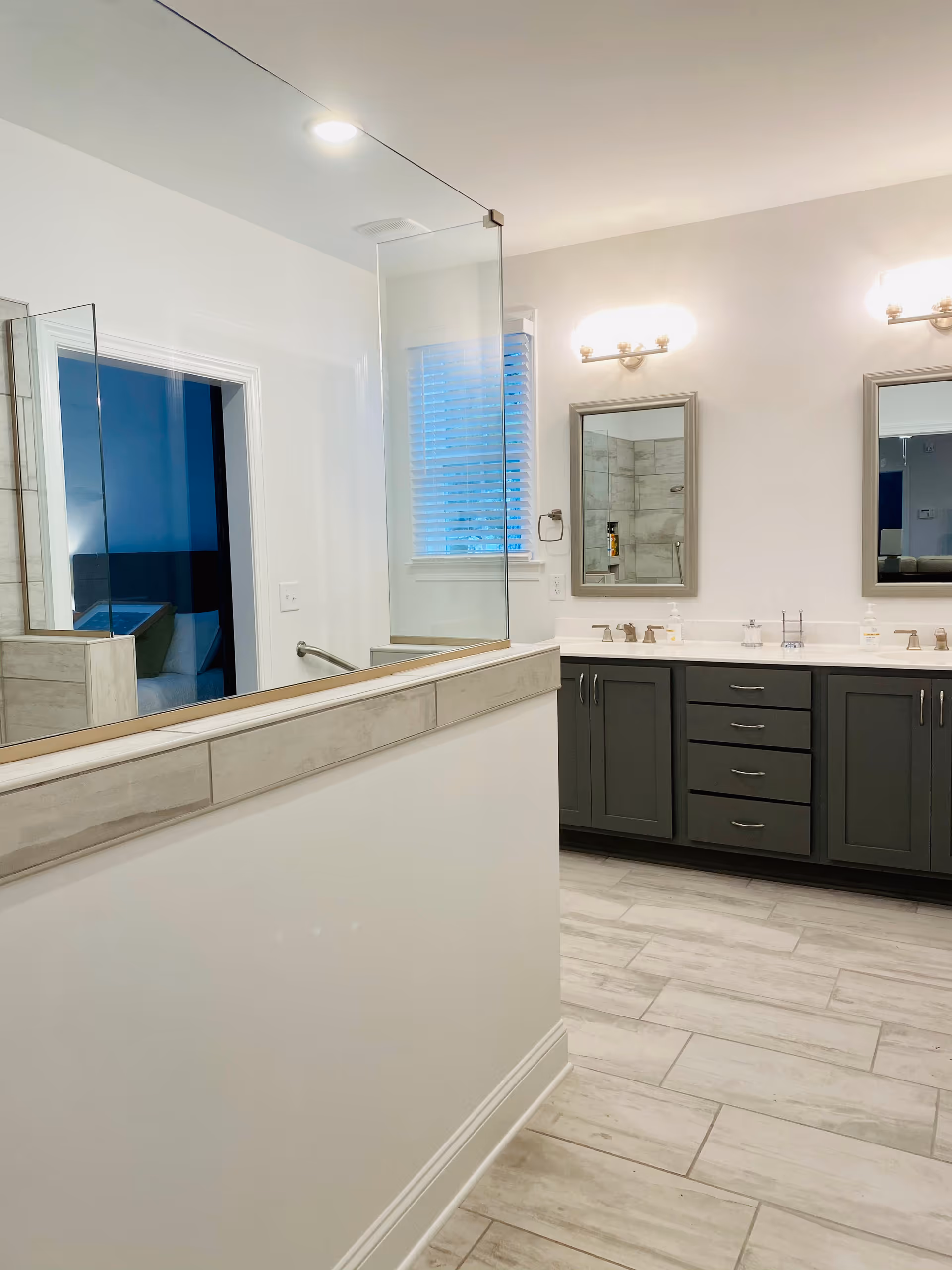 A modern bathroom with a large mirror above a double sink vanity with dark cabinets. Two wall-mounted light fixtures are above the mirrors. There is a window with blinds and a glass shower enclosure visible in the reflection.