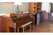 Upright wooden piano with a bench in a communal living area with hardwood floors, shelving, and a small kitchenette in the background.