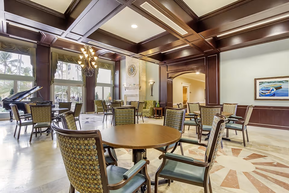 A spacious and elegant common area with multiple round tables and chairs arranged on a polished marble floor. The room features large windows letting in natural light, a grand piano near the windows, a decorative fireplace, and a coffered ceiling with dark wood beams. There is a painting on the wall and a doorway leading to another room.