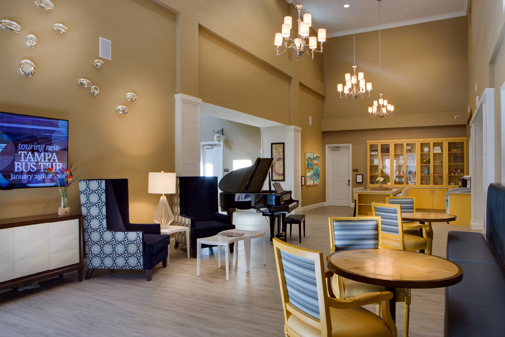 A spacious and well-lit common area in a senior living facility featuring a grand piano, comfortable armchairs, round wooden tables with yellow and blue striped chairs, a wall-mounted TV displaying information about a Tampa bus trip, modern chandeliers, and a yellow cabinet with decorative items.