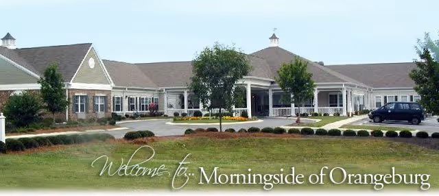 Front exterior view of a single-story senior living facility building with a driveway, landscaped lawn, and trees. A vehicle is parked near the entrance, and the text 'Welcome to Morningside of Orangeburg' is overlaid on the image.