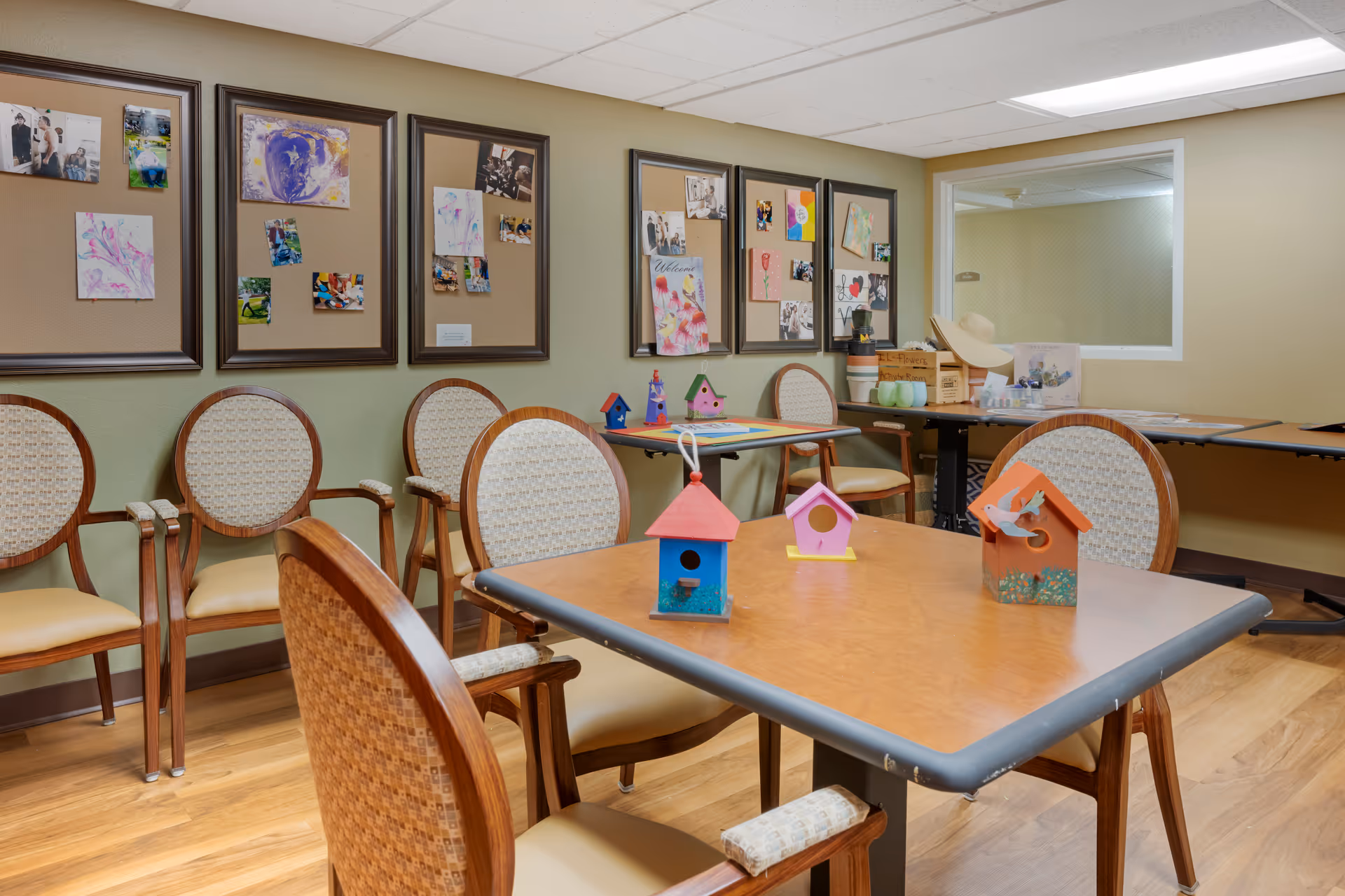A room with several wooden chairs arranged around tables. On the tables are colorful small birdhouse crafts. The walls have framed bulletin boards displaying photos and artwork. The floor is wooden, and there is a window with a wire mesh looking into another room.
