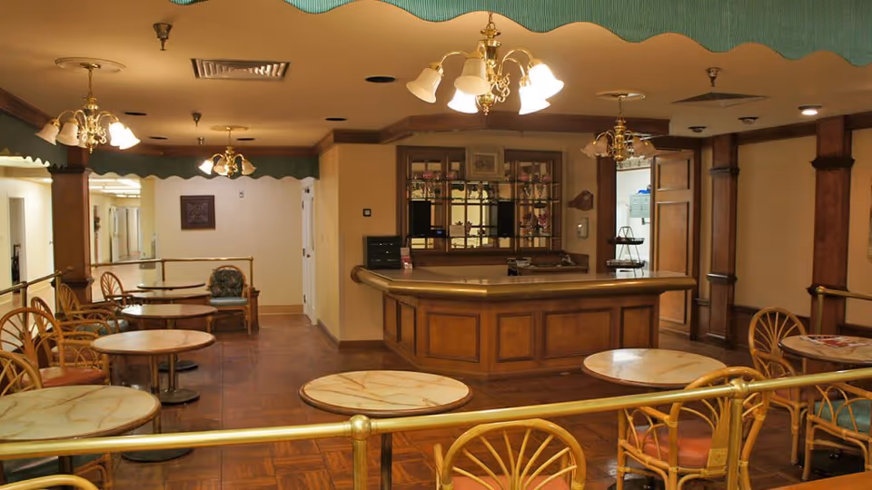 Interior view of a common area in an assisted living facility featuring several round tables with chairs arranged around them. The room has warm lighting with multiple ceiling light fixtures and wooden accents. There is a wooden counter or reception desk at the back with a glass display cabinet behind it. The floor is tiled and there are brass railings separating the seating area from the walkway.