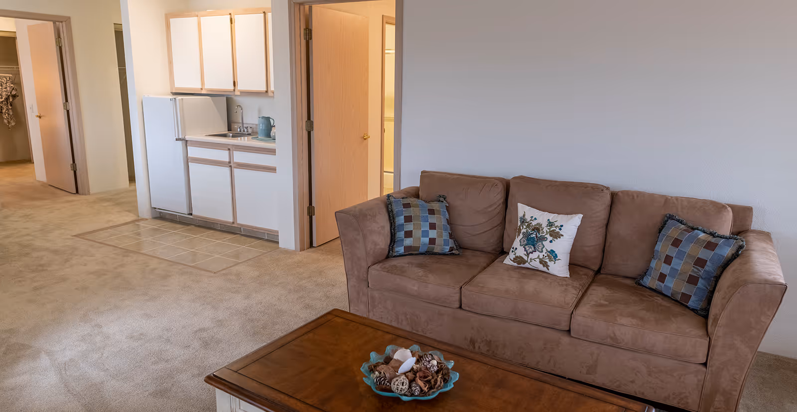 Interior view of a senior living apartment showing a brown sofa with three decorative pillows, a wooden coffee table with a decorative bowl, a small kitchenette with white cabinets and a refrigerator, and open doors leading to other rooms.