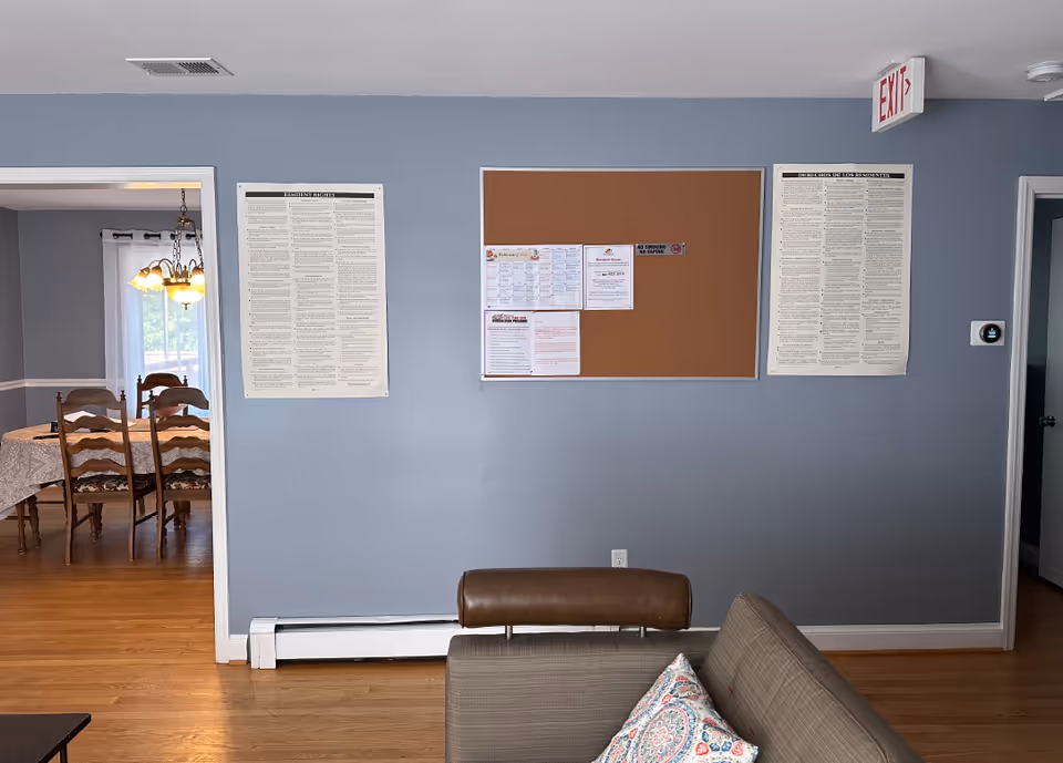 Interior view of a senior living facility room with a gray wall featuring a bulletin board and two large posters. To the left, there is an open doorway leading to a dining room with a wooden table and chairs under a chandelier. In the foreground, part of a sofa with a patterned pillow is visible. The floor is wooden, and an exit sign is mounted on the ceiling near the right doorway.