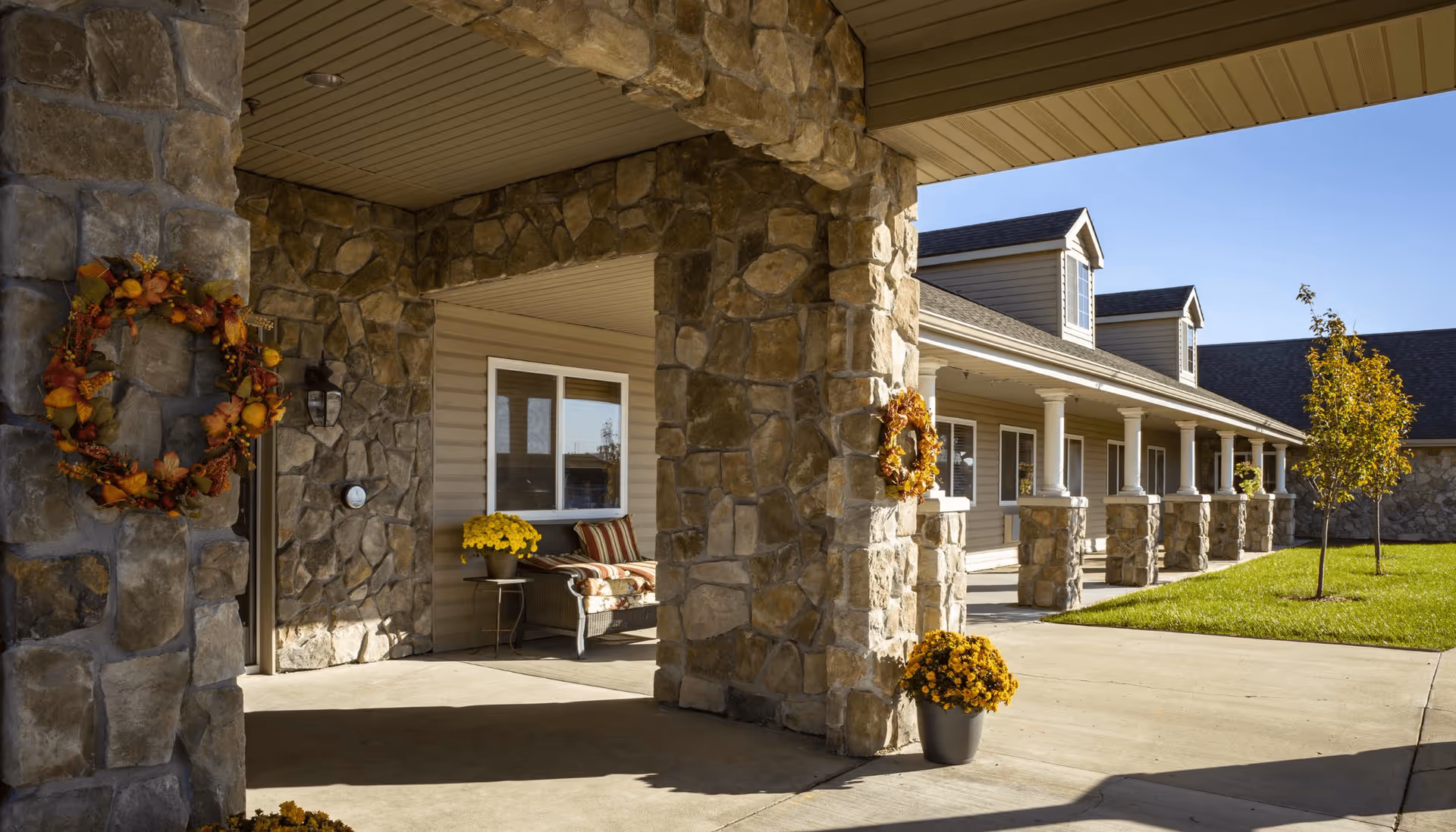 Exterior view of a senior living facility entrance with stone pillars decorated with autumn wreaths and potted yellow flowers. There is a covered porch area with a bench and cushions, and a well-maintained lawn with a small tree under a clear blue sky.