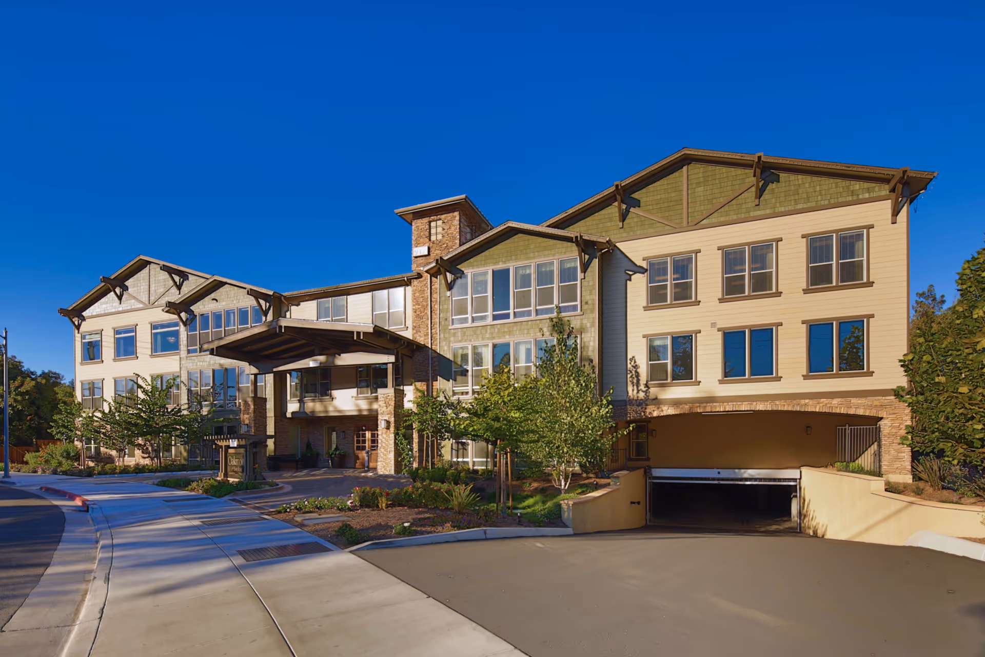 Exterior view of a multi-story senior living facility building with beige and green siding, large windows, a covered entrance, and an underground parking garage entrance. The building is surrounded by landscaped greenery and trees under a clear blue sky.
