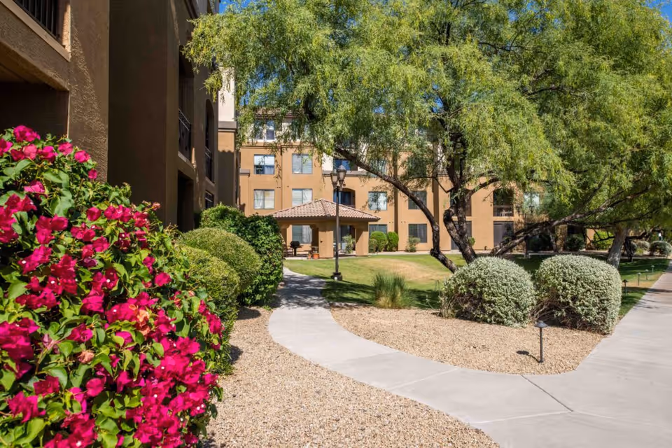 Outdoor garden area at La Siena facility with a paved walkway, green trees, bushes, and vibrant pink flowers. Beige multi-story building with balconies is visible in the background under a clear blue sky.