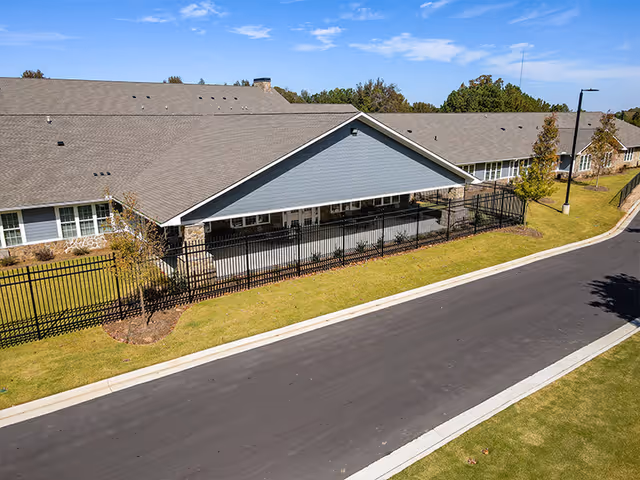 Exterior view of a single-story assisted living facility building with a long roof, stone and siding walls, black metal fencing, a paved road, and green lawns under a blue sky with some clouds.