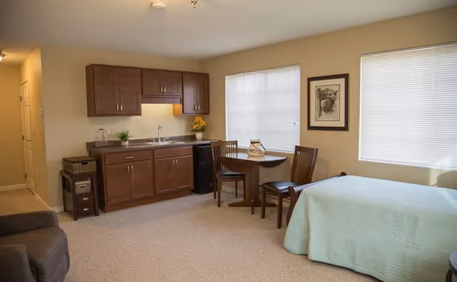 A senior living facility room with a small kitchenette featuring wooden cabinets, a sink, and a mini fridge. There is a round dining table with two chairs next to two windows with blinds. A bed with a light green bedspread is visible on the right side, and a comfortable armchair is partially visible on the left. The walls are beige, and there is a framed picture hanging between the windows.