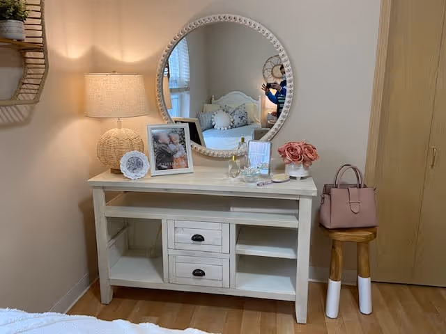 A cozy bedroom corner featuring a white wooden dresser with two drawers and open shelves. On top of the dresser is a round mirror with a decorative frame, a table lamp with a woven base, framed photos, a glass of white wine, and a small vase of pink flowers. To the right of the dresser is a wooden stool with white painted legs holding a pink handbag. The reflection in the mirror shows a bed with white bedding and decorative pillows.
