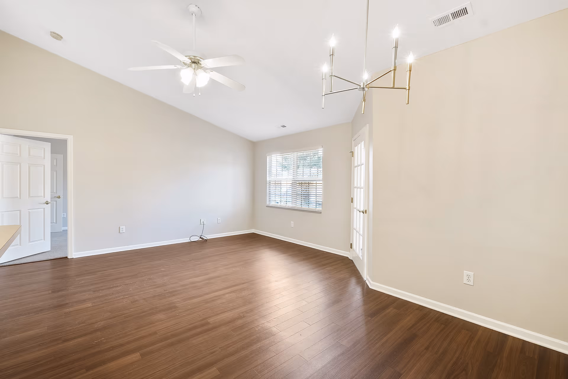 Empty room with light beige walls, dark wood flooring, a ceiling fan with lights, a modern chandelier, a window with blinds, a white door leading to another room, and a glass door on the right side.