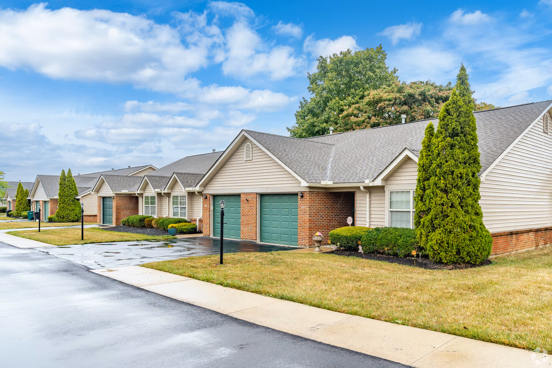 Row of single-story residential units with attached garages, green garage doors, and well-maintained lawns under a partly cloudy blue sky.