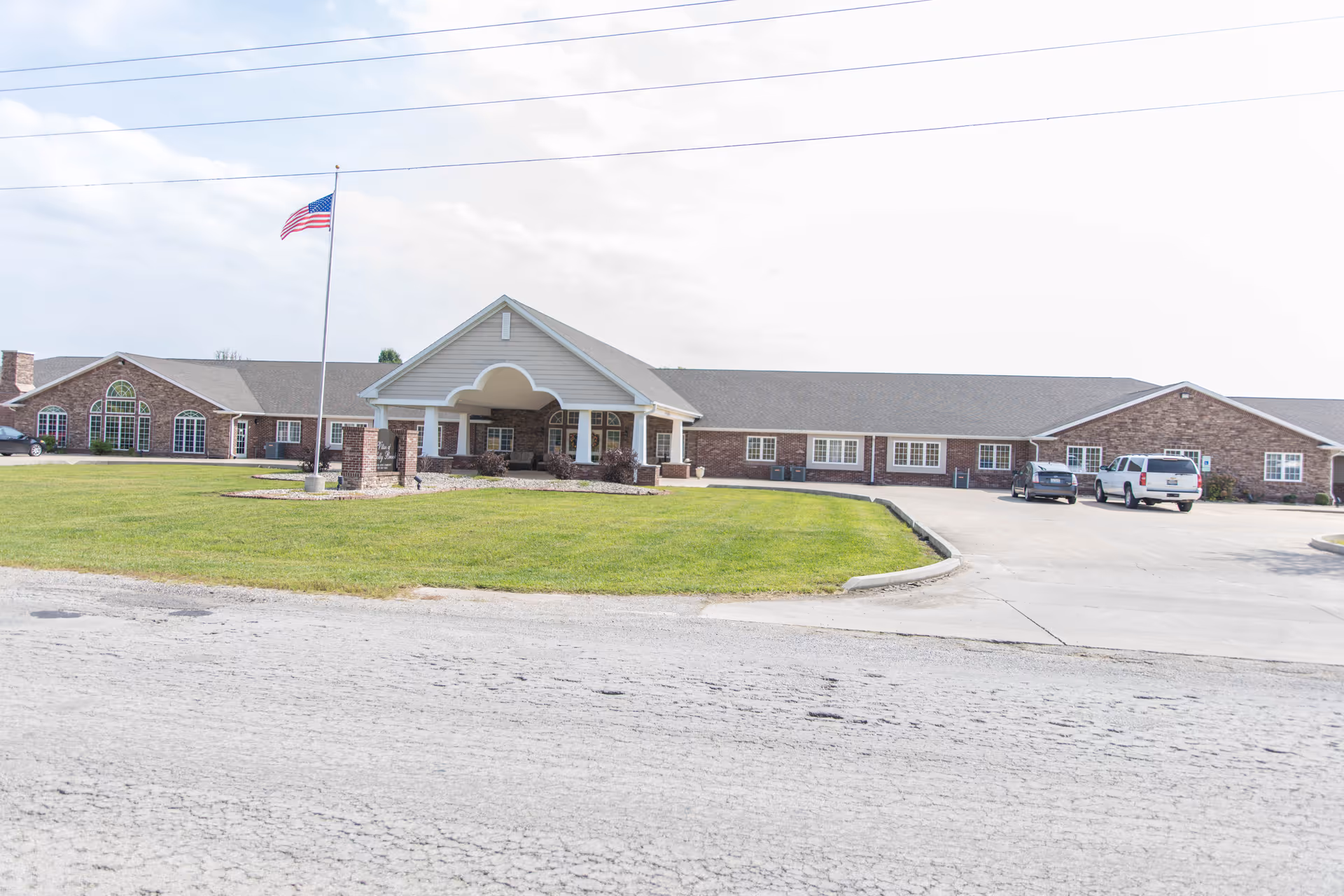 One-story brick senior living building with a covered porte-cochere entrance, flagpole, grassy lawn, and a parking area.