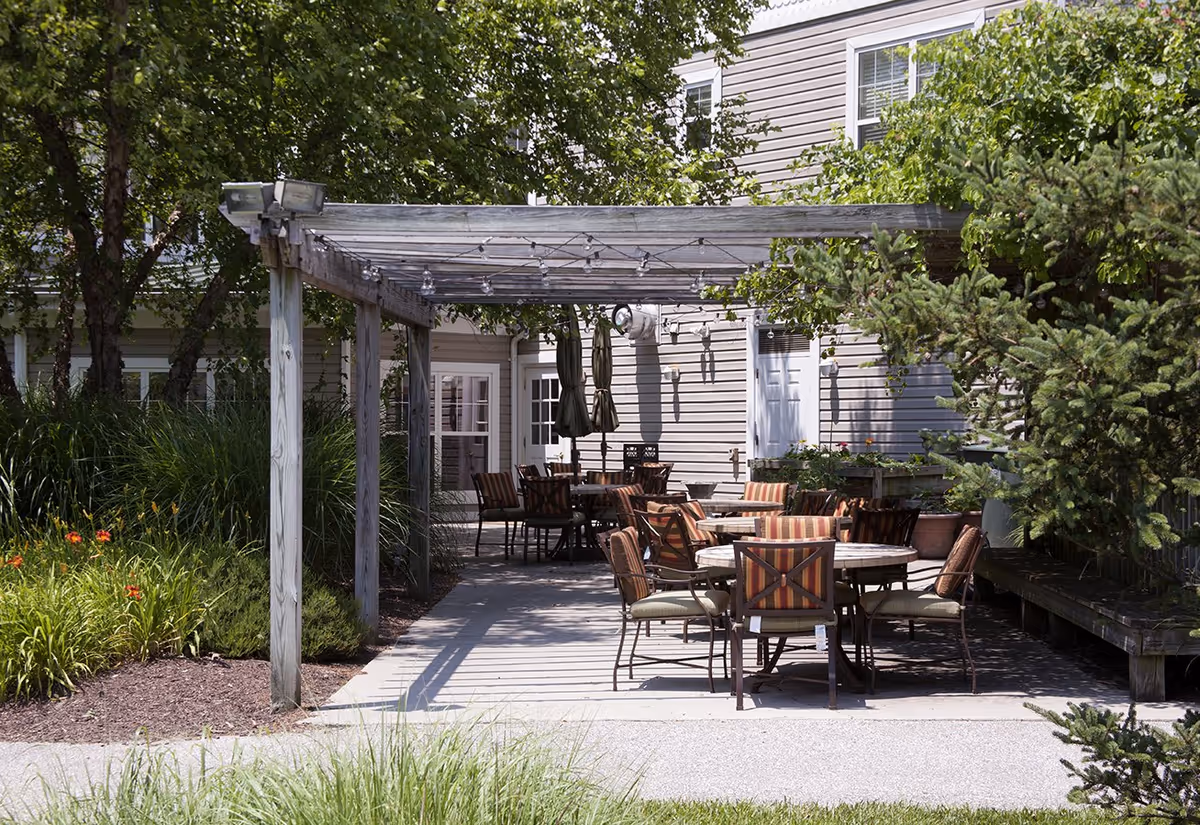 Outdoor patio area with several round tables and cushioned chairs under a wooden pergola attached to a building. The patio is surrounded by greenery including trees, bushes, and flowering plants.