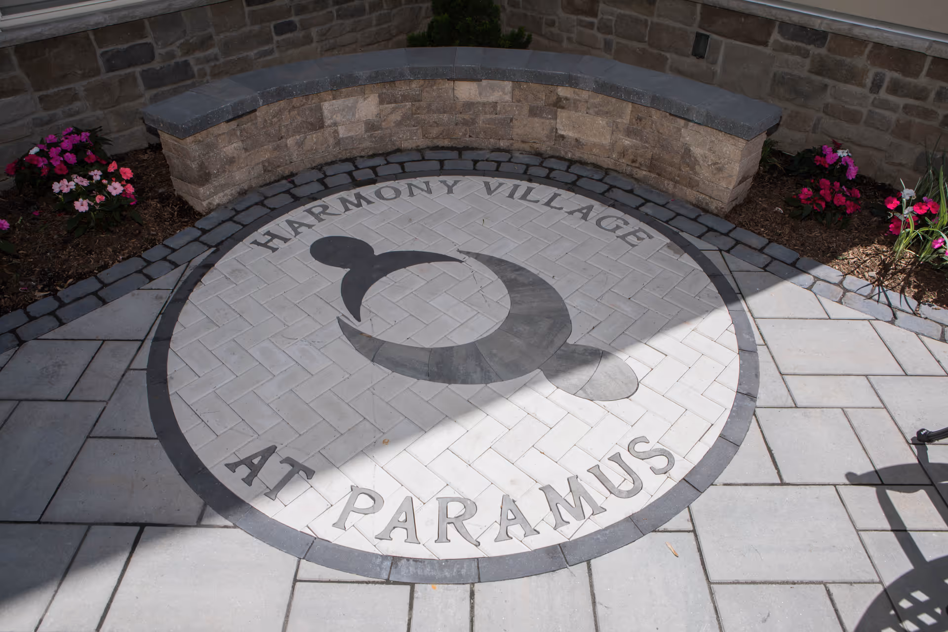 Outdoor stone patio area with a circular stone inlay on the ground featuring the text 'Harmony Village at Paramus' and a logo of two abstract human figures holding hands. There is a curved stone bench against a brick wall and flower beds with pink and red flowers on either side.
