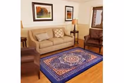 A cozy living room with a beige sofa, two brown armchairs, a wooden side table with a lamp, and a blue patterned area rug on a wooden floor. Two framed pictures hang on the wall above the sofa.
