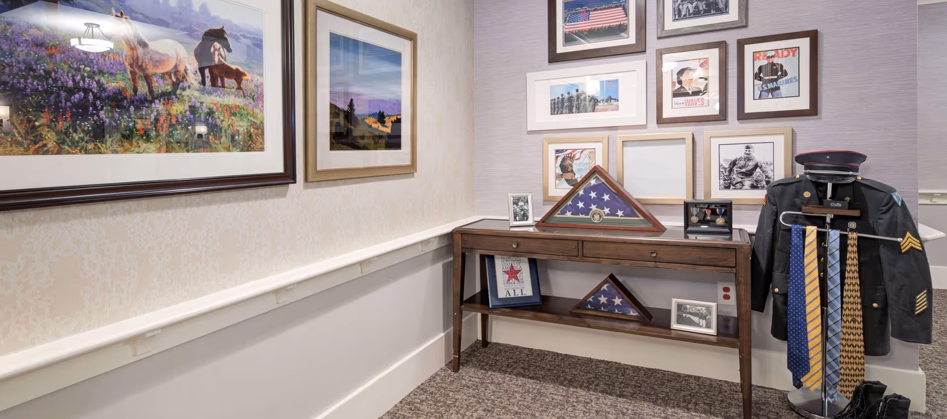 A corner of a room featuring a wooden console table displaying folded American flags in triangular cases, framed photographs, and military medals. To the right, a military uniform jacket is displayed on a stand with several ties hanging from it. The wall behind the table is decorated with framed patriotic and military-themed pictures. On the left wall, there are two large framed paintings, one depicting horses in a field of flowers.