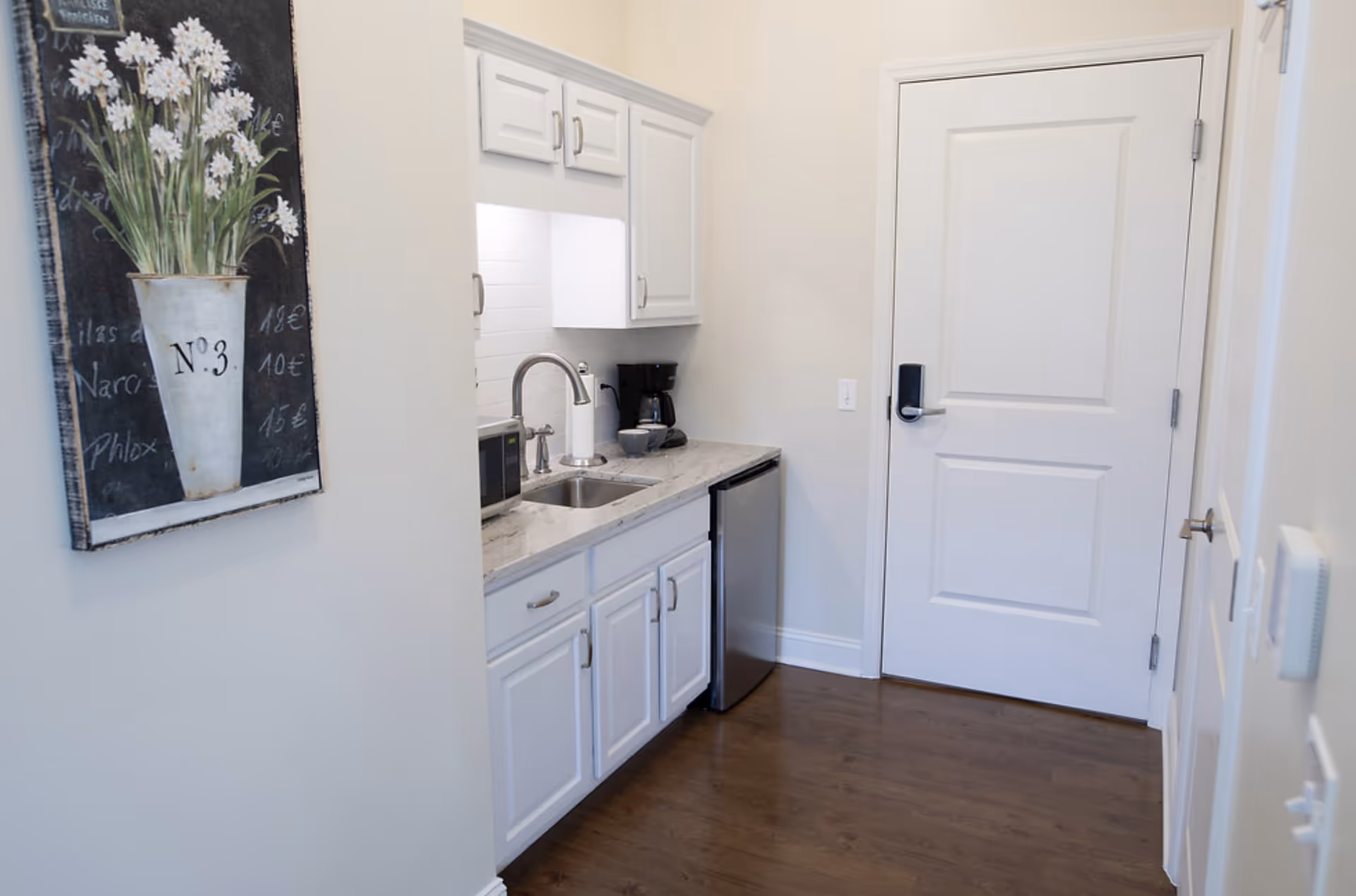 Small kitchenette with white cabinets, a sink, microwave, mini fridge and coffee maker next to a closed white door and floral wall art.