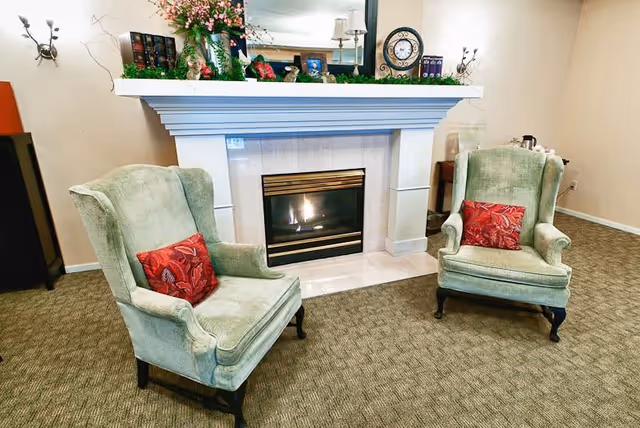A cozy interior space featuring a lit fireplace with a white mantel decorated with flowers, books, and small framed pictures. In front of the fireplace are two green upholstered armchairs, each with a red decorative pillow. The room has beige walls and carpeted flooring.