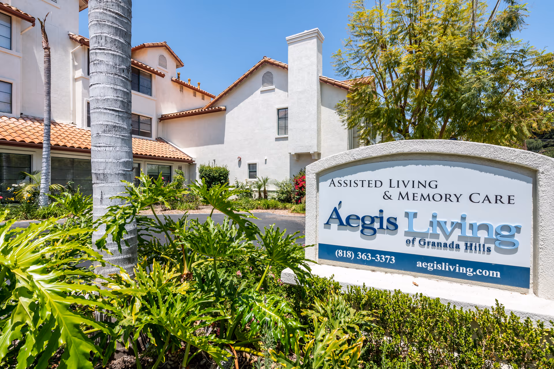 Exterior view of Aegis Living Granada Hills assisted living and memory care facility with a white building featuring terracotta roof tiles, surrounded by green plants and palm trees under a clear blue sky. A large sign in front displays the facility name, phone number, and website.