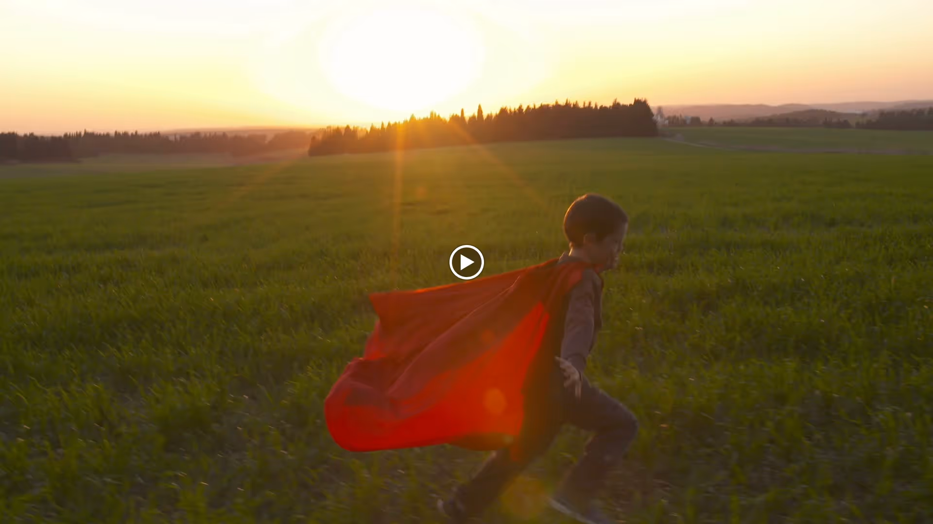 A young child wearing a red cape runs through a green field at sunset with the sun low on the horizon and trees in the background.
