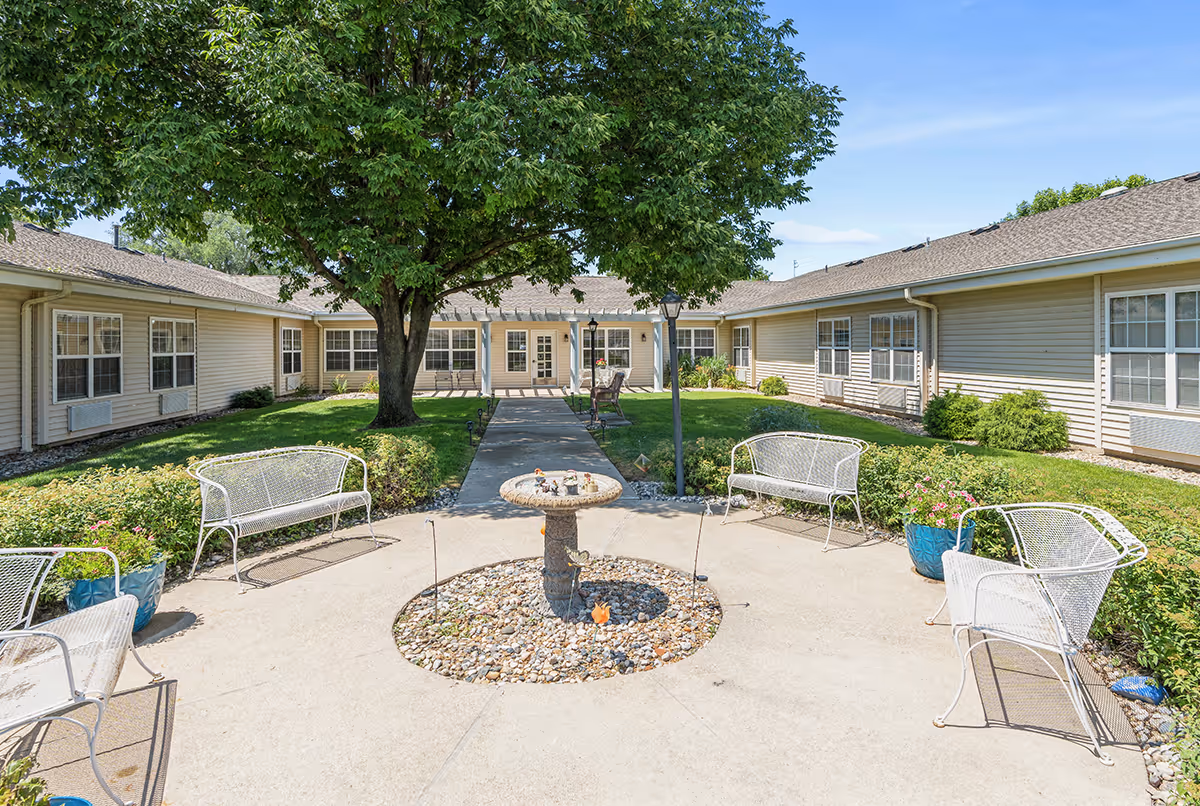 Outdoor courtyard area at Floyd Senior Living with a large tree in the center, surrounded by a paved walkway and white metal benches. The courtyard is bordered by a single-story building with multiple windows and a clear blue sky overhead.